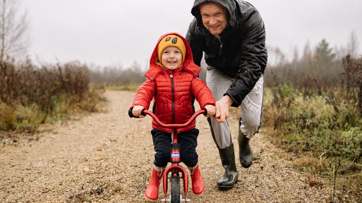 A father joyfully helps his child ride a bicycle on a gravel path in an autumn setting.